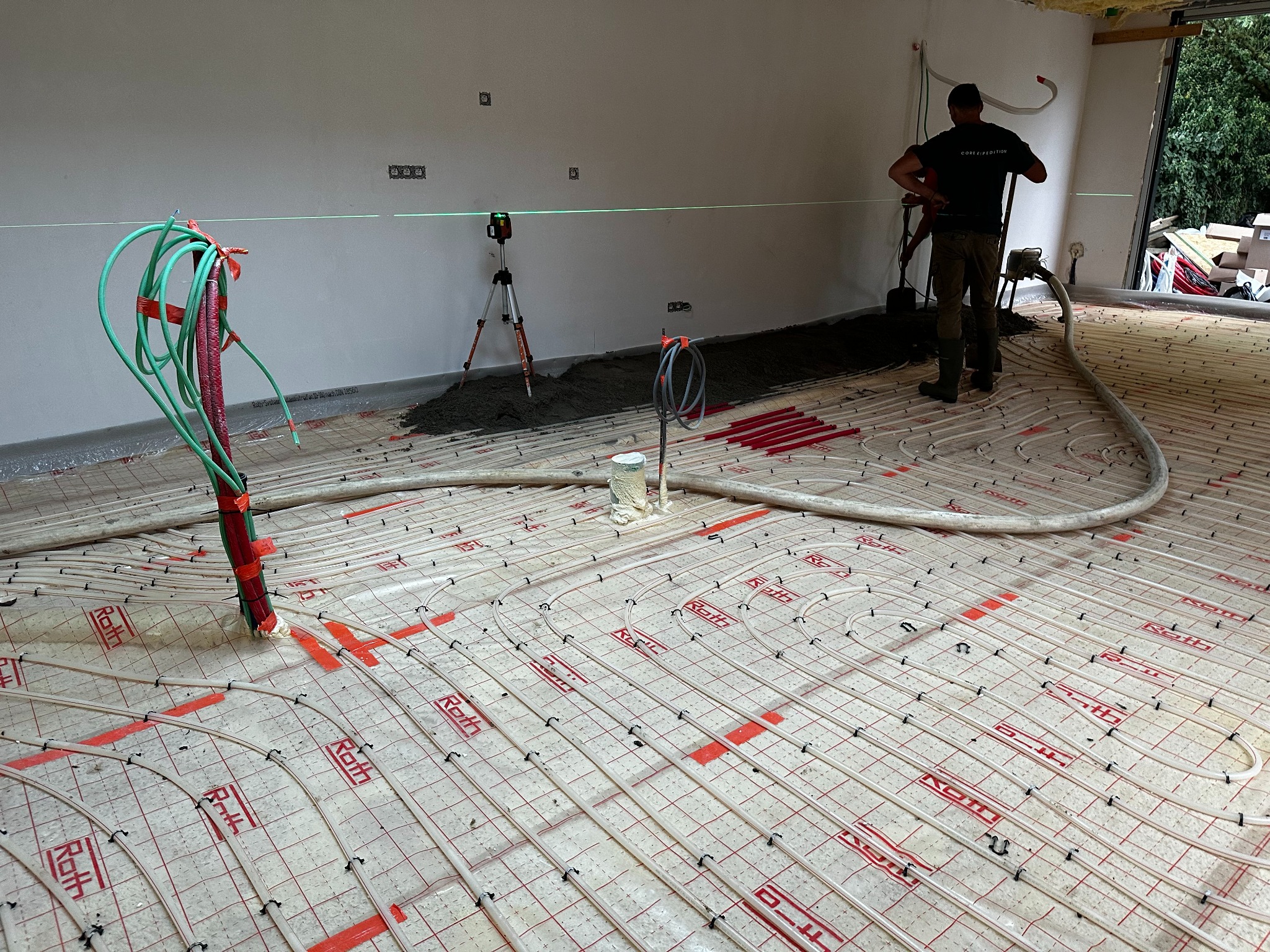 Układanie ogrzewania podłogowego Roth z użyciem poziomicy laserowej. Widoczny pracownik w trakcie wylewania posadzki. Rury grzewcze zamocowane do podłoża.