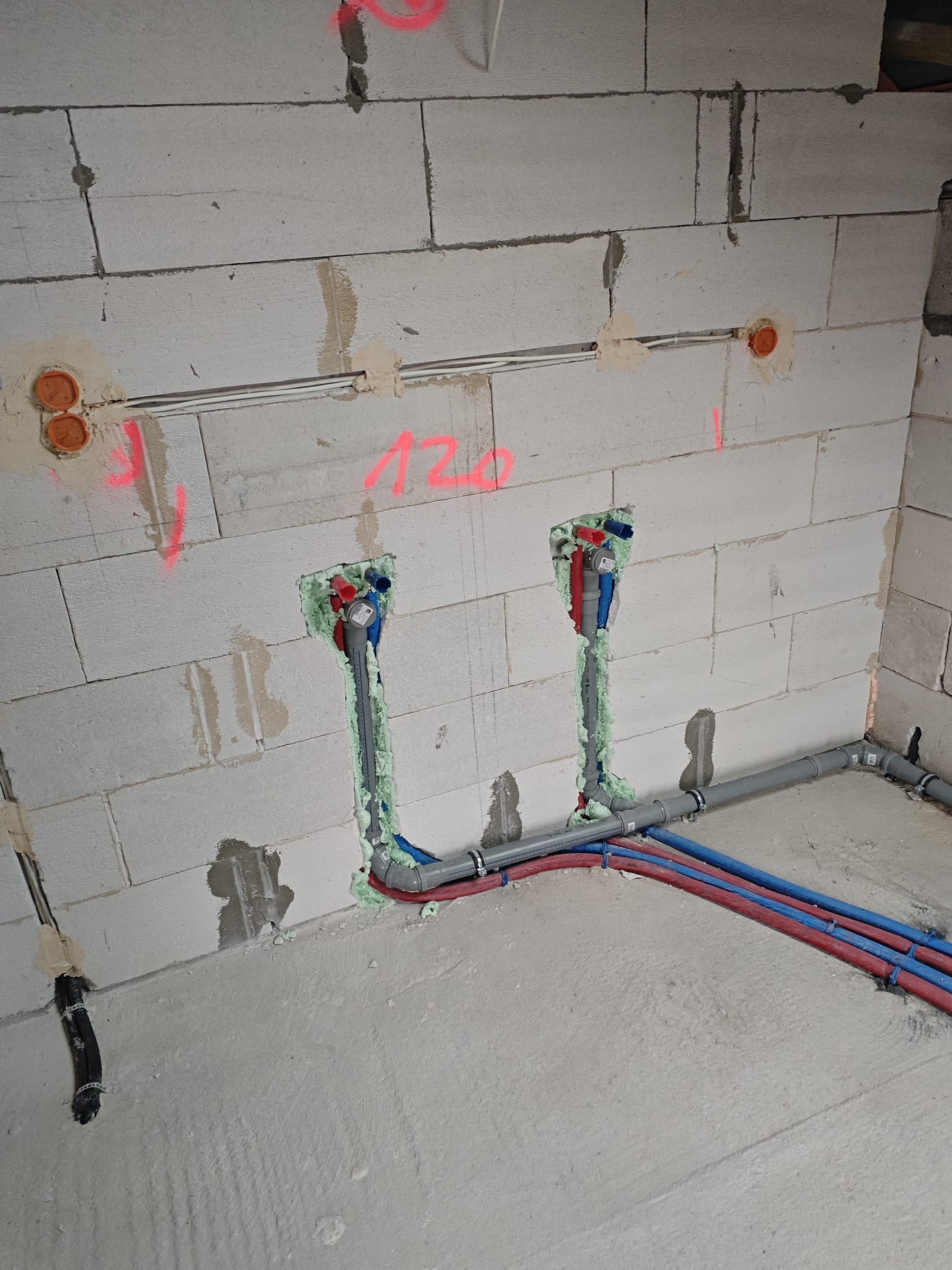 Surowa ściana z betonu komórkowego z instalacją wodną w trakcie montażu, rury czerwone i niebieskie, pianka montażowa, widoczne przewody elektryczne.