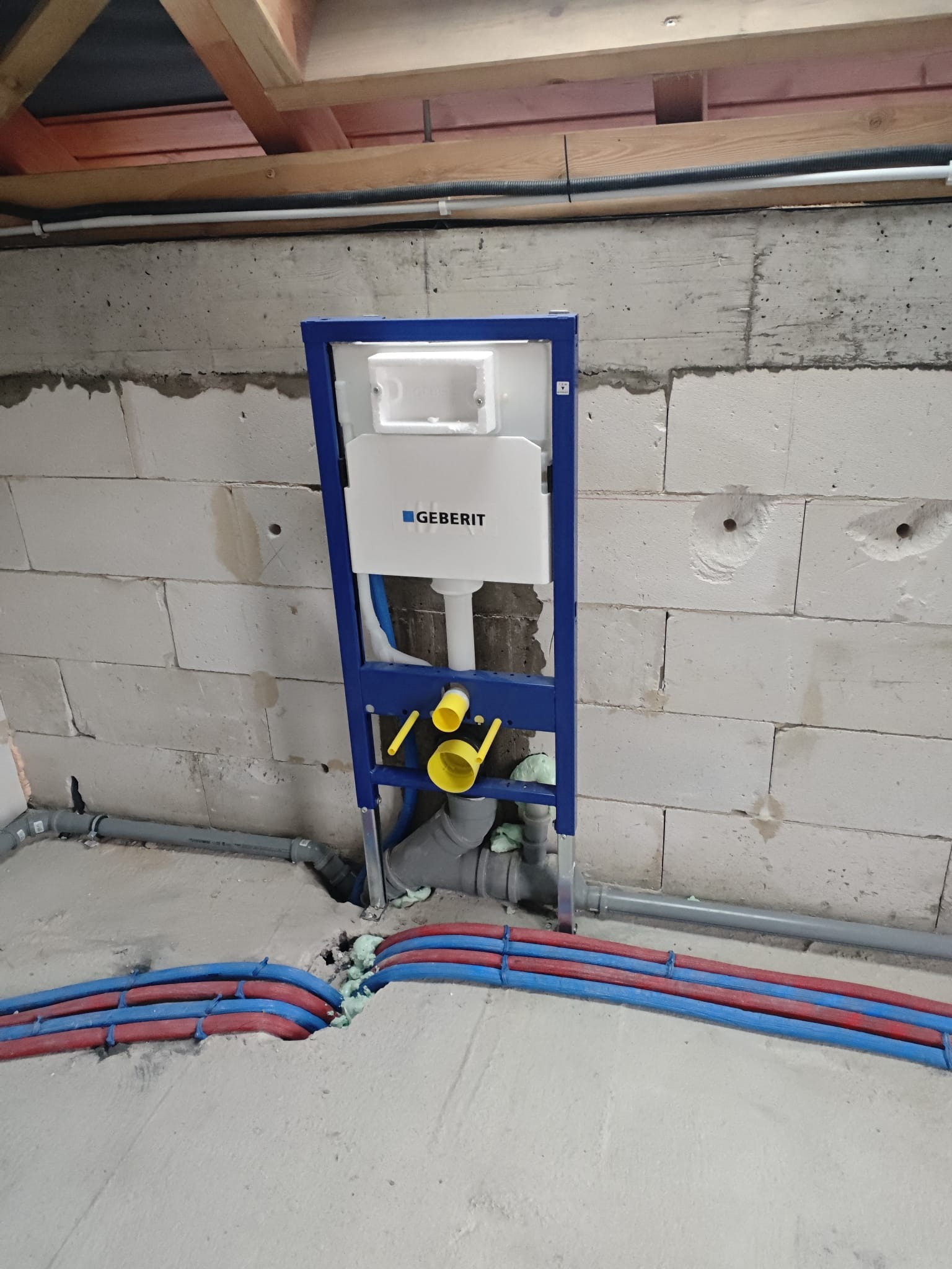 Stelaż podtynkowy Geberit zamontowany przy ścianie z betonu komórkowego, widoczne rury instalacji wodnej w kolorze czerwonym i niebieskim, szare rury kanalizacyjne.