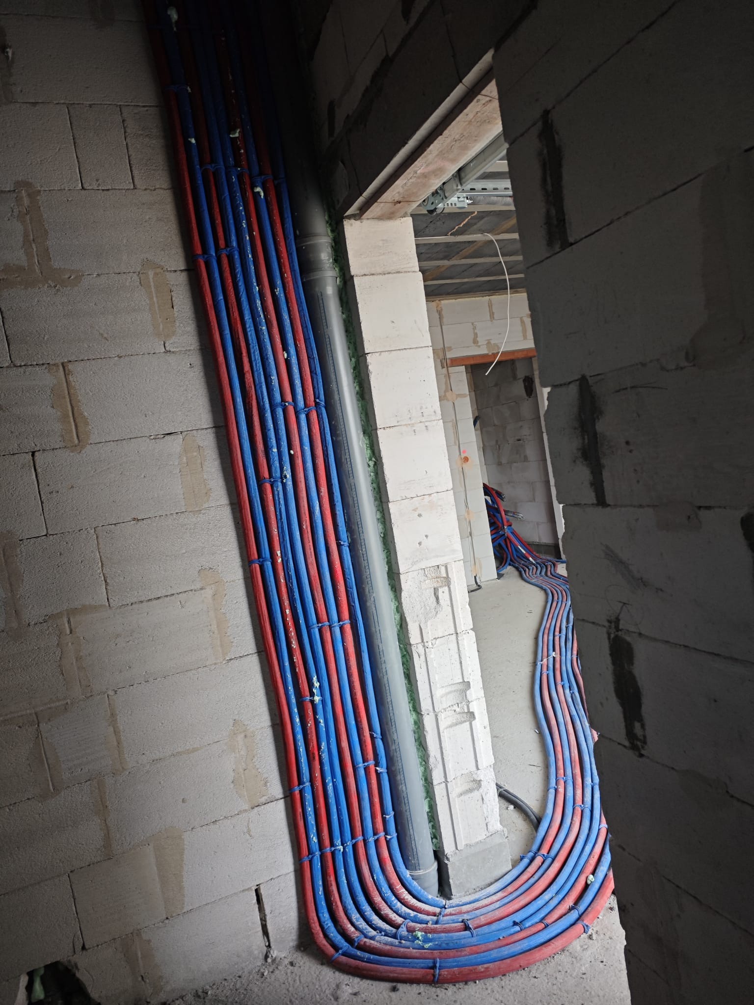 Instalacja wodna z rur PEX w kolorach czerwonym i niebieskim, prowadzona wzdłuż ściany z betonu komórkowego, widok z perspektywy korytarza w surowym stanie.