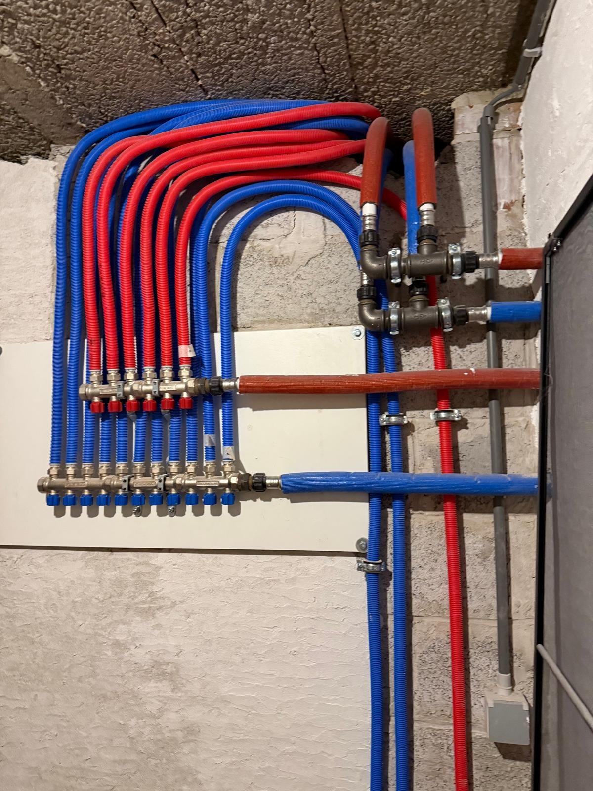 Rozdzielacz instalacji CO i CWU z rurami w kolorze niebieskim i czerwonym, zamontowany na ścianie z surowego betonu. Widoczne liczne złączki i zawory.