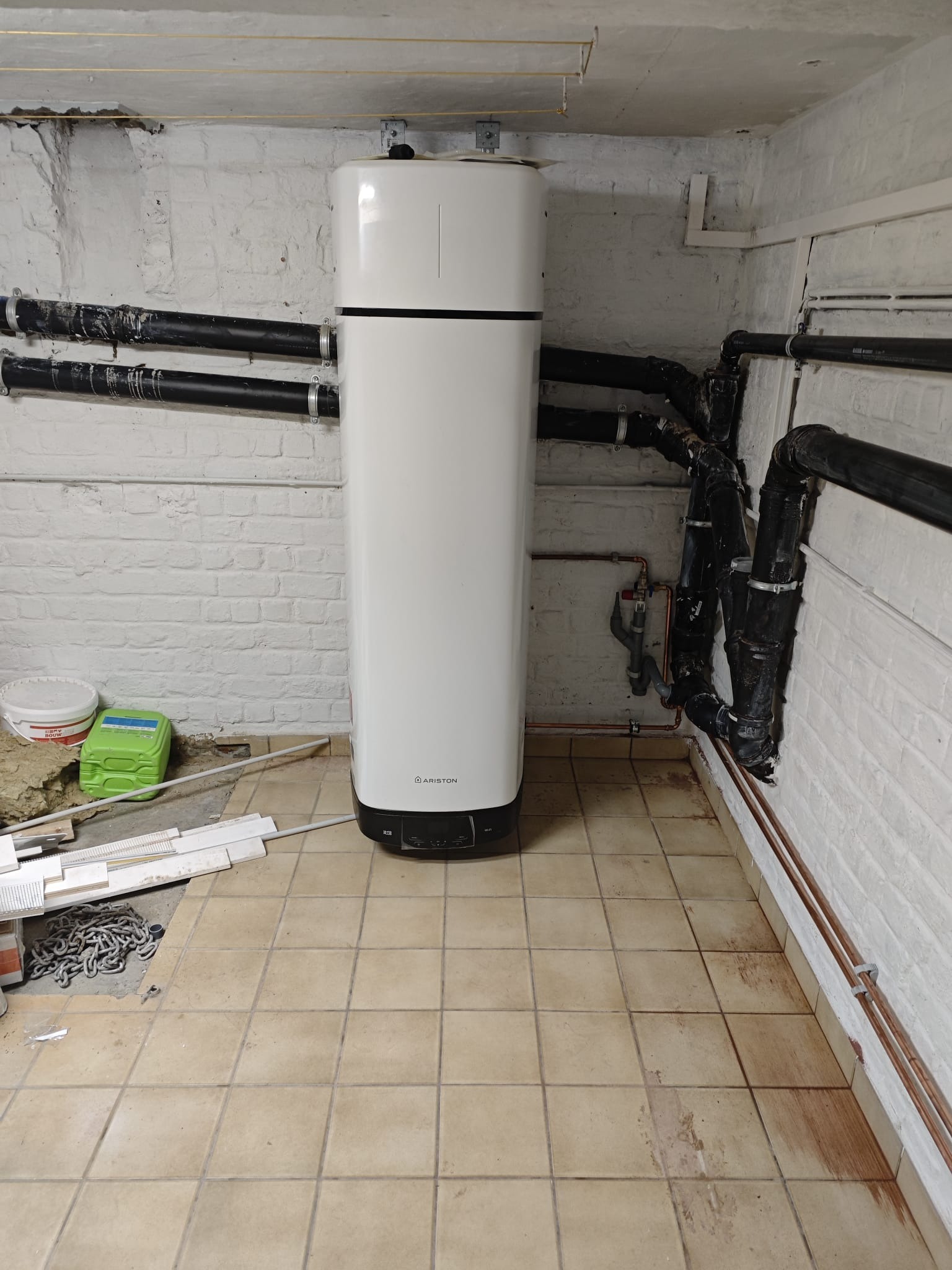 Biały, nowoczesny bojler Ariston w kotłowni z instalacją. Widoczne rury, ceglana ściana i płytki podłogowe. Ujęcie z dołu, podkreślające wysokość urządzenia.