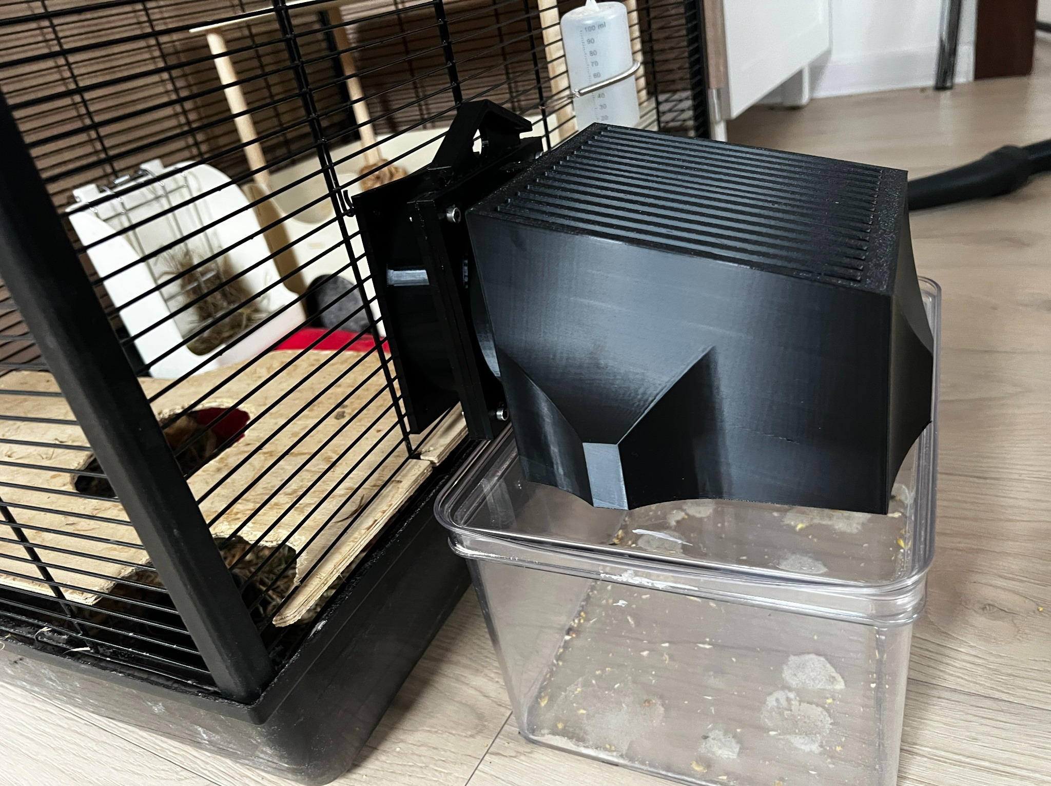 Czarny, plastikowy podajnik na karmę, przymocowany do klatki dla zwierząt, z przezroczystym pojemnikiem na dnie. Widoczne ślady druku 3D.