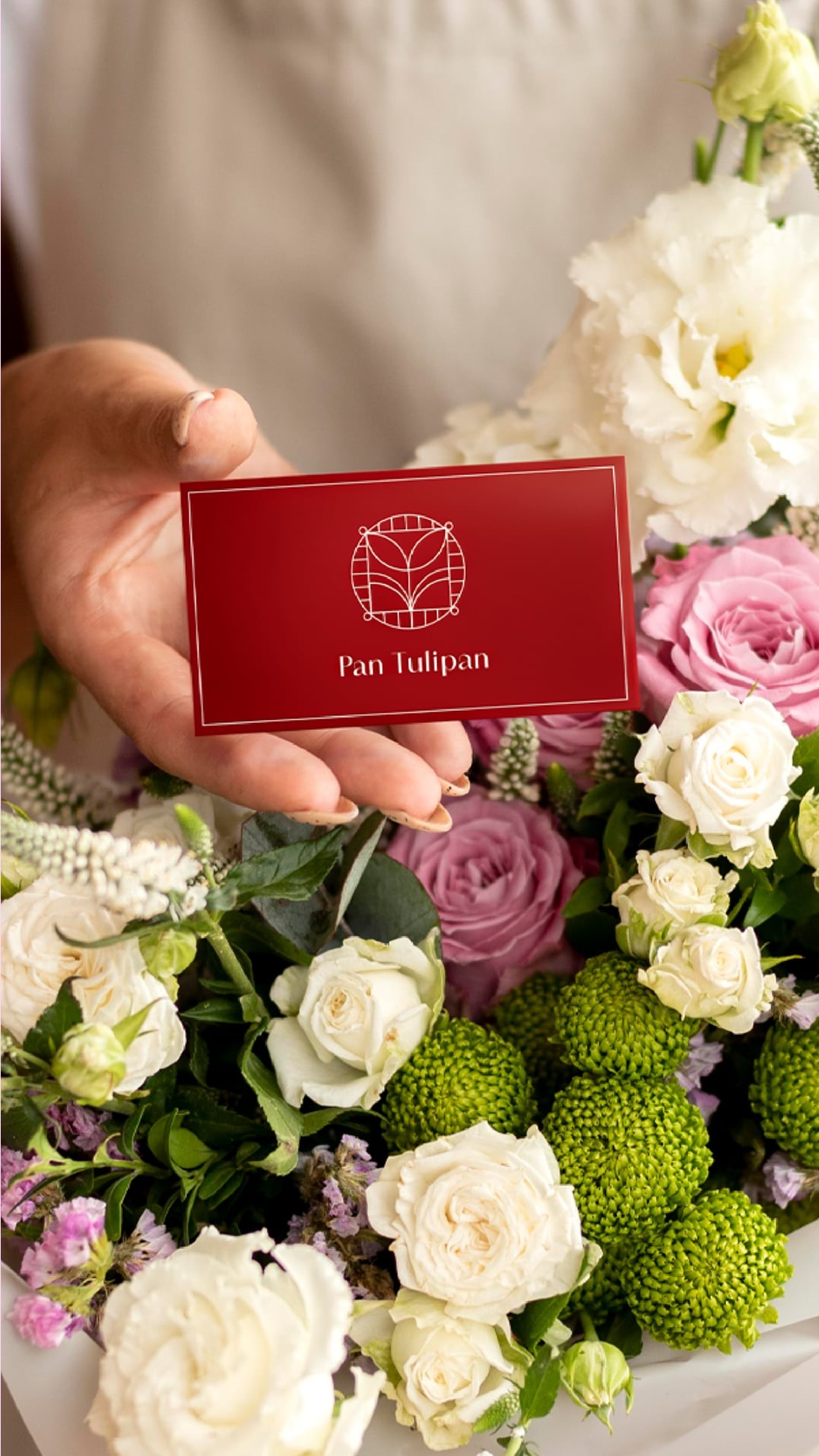 Czerwona wizytówka z białym logo 'Pan Tulipan' trzymana nad bukietem kwiatów w odcieniach bieli, różu i zieleni. Delikatne oświetlenie, elegancki minimalizm.