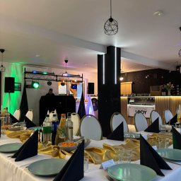 ADbiuro Dorota Adamowska - Uroczysty stół z dekoracjami w odcieniach złota i czerni, przygotowany na imprezę. W tle widoczny DJ i oświetlenie sceniczne, tworzące imprezowy klimat.