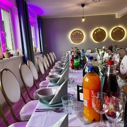 ADbiuro Dorota Adamowska - Uroczysty stół z zastawą, napojami i dekoracjami kwiatowymi, przygotowany na przyjęcie w jasnym wnętrzu z oświetleniem punktowym i fioletowymi akcentami.