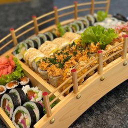 ADbiuro Dorota Adamowska - Zestaw sushi, różnorodne rolki ułożone na drewnianej tacy w kształcie mostu, z dekoracjami z imbiru i sałaty, prezentacja dań kuchni japońskiej.