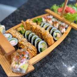 ADbiuro Dorota Adamowska - Zestaw sushi w drewnianej łódce: różnorodne rolki z krewetkami w tempurze, łososiem i warzywami, udekorowane sosem i sezamem, podane na ciemnym blacie.