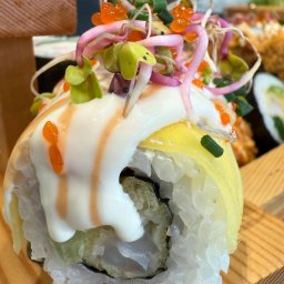 ADbiuro Dorota Adamowska - Sushi rolka z omletem, kremowym sosem i kiełkami, podana na drewnianej desce. Widoczne inne rolki w tle. Ujęcie z bliska, podkreślające detale i świeżość składników.