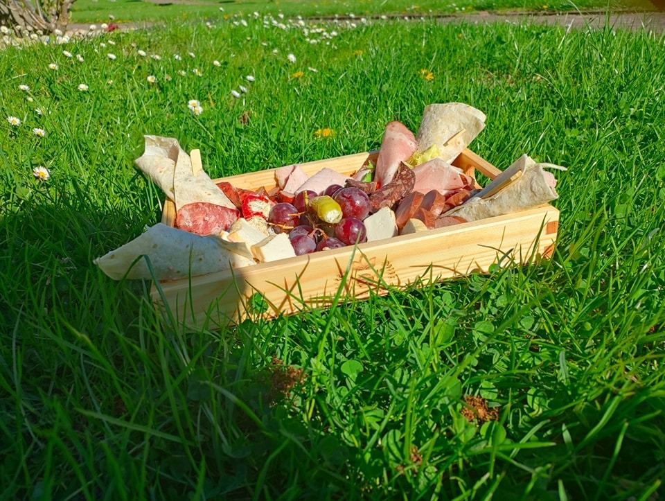 Drewniana skrzynka z przekąskami (sery, wędliny, owoce, tortille) na trawie w plenerze, w słoneczny dzień. Idealne na piknik lub imprezę.