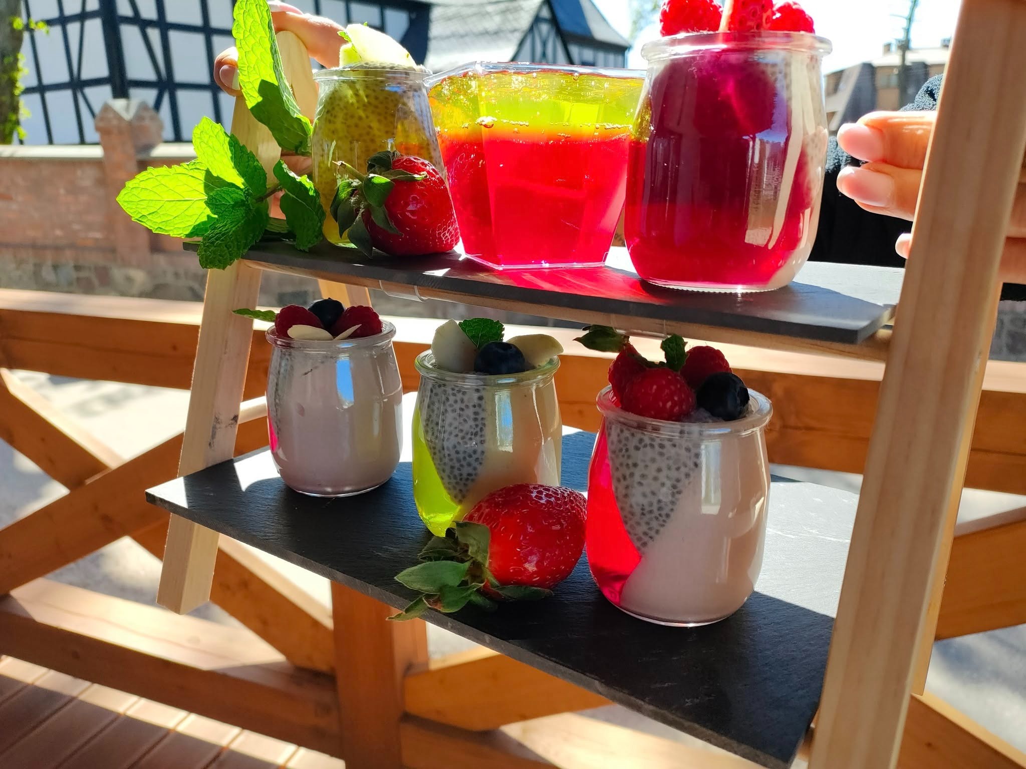 Kolorowe desery w szklanych naczyniach, udekorowane owocami i miętą, ustawione na drewnianym stojaku. Słoneczne światło podkreśla świeżość składników.