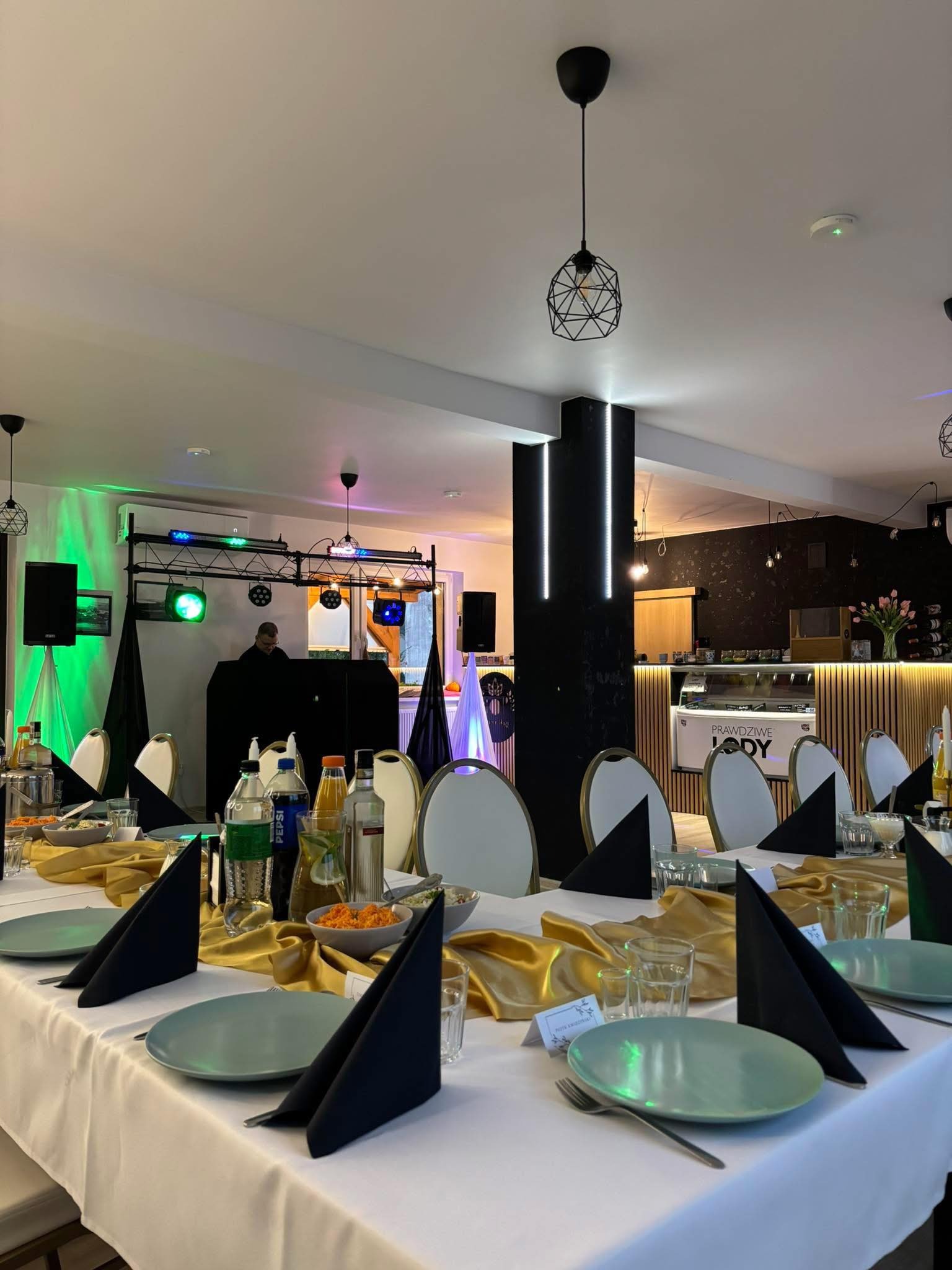 Uroczysty stół z dekoracjami w odcieniach złota i czerni, przygotowany na imprezę. W tle widoczny DJ i oświetlenie sceniczne, tworzące imprezowy klimat.