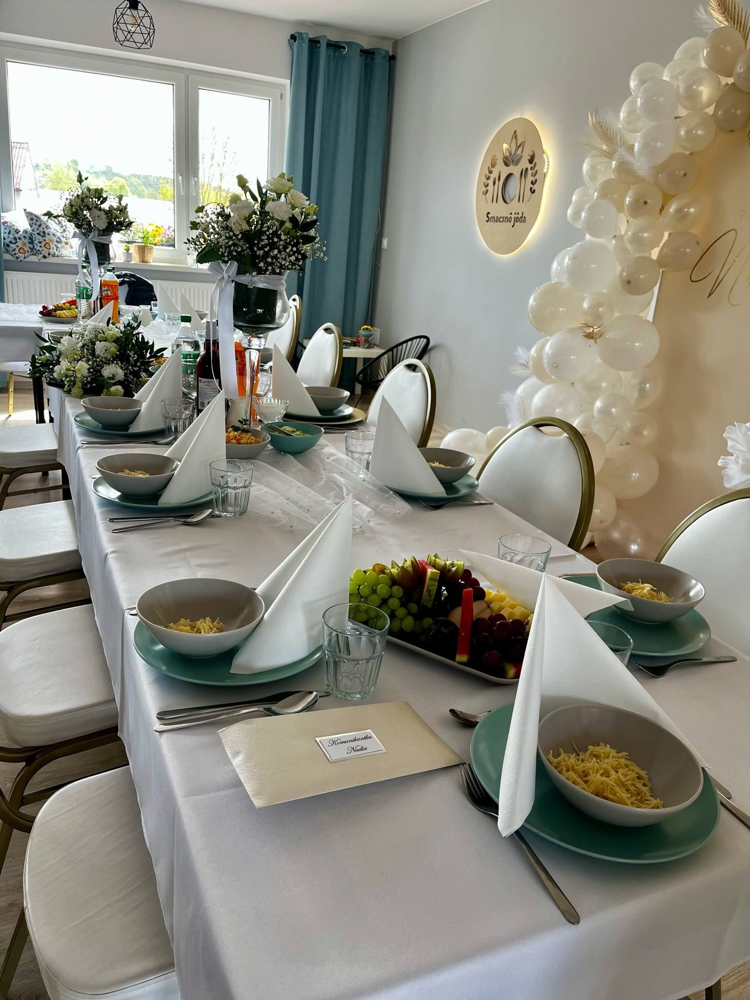 Elegancko nakryty stół z dekoracjami kwiatowymi i balonowymi, przygotowany na uroczystość. Widoczne talerze z potrawami, owoce i wizytówki gości.