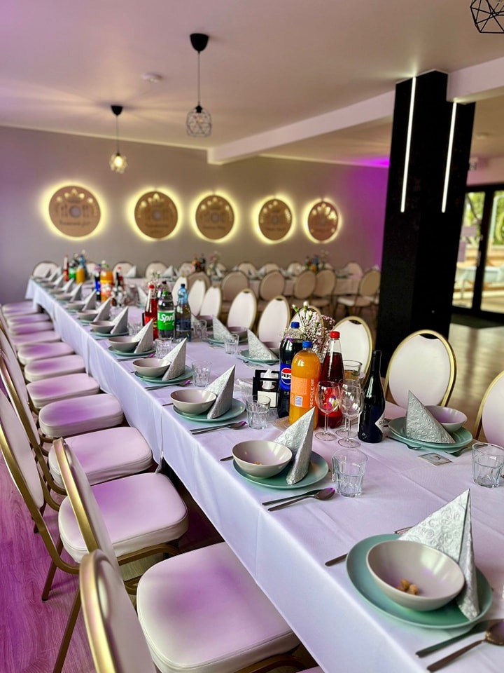 Uroczysty stół bankietowy z białym obrusem, nakryciami w odcieniach zieleni i napojami, przygotowany na przyjęcie w sali z dekoracyjnym oświetleniem.