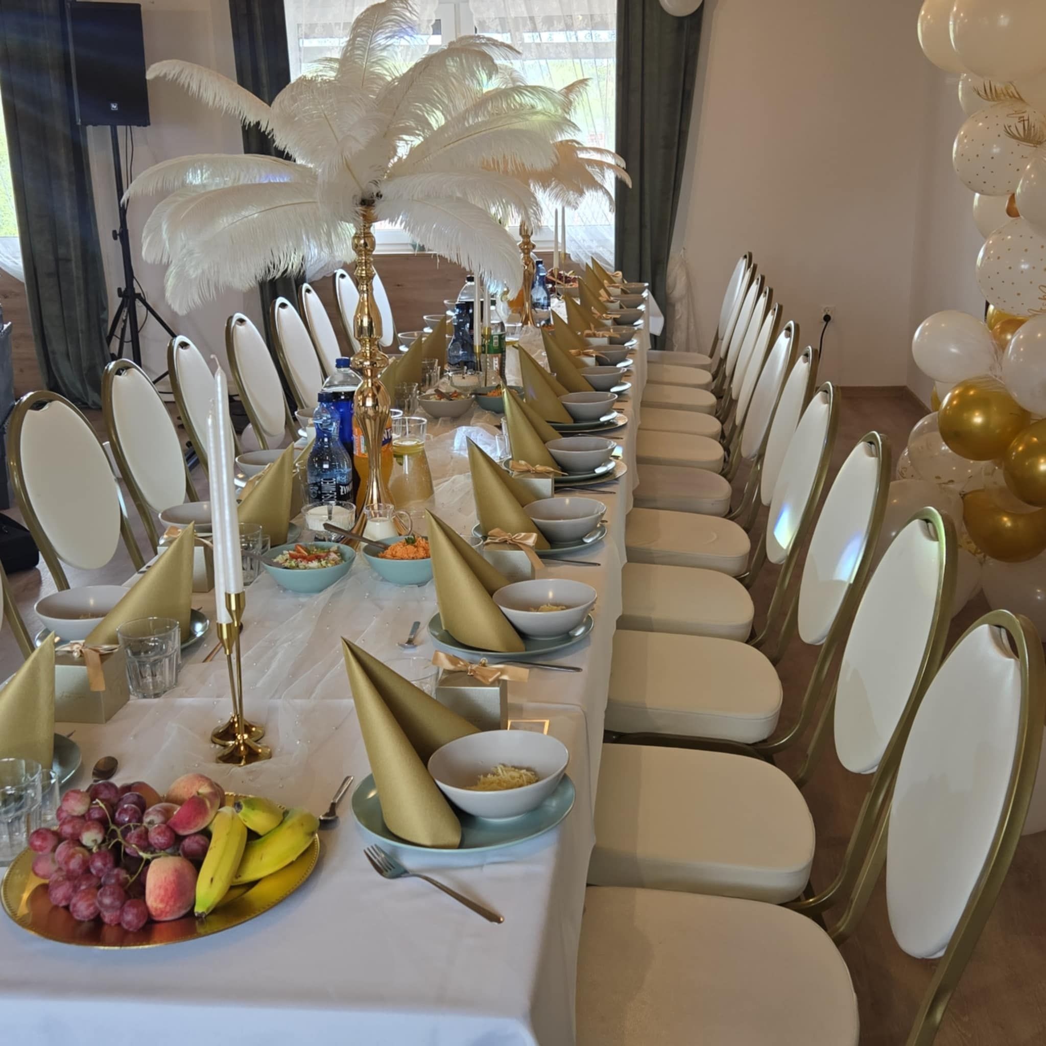 Elegancko nakryty stół na przyjęcie z białym obrusem, złotymi serwetami, owocami i dekoracjami z piór. W tle balony i krzesła w stylu glamour.
