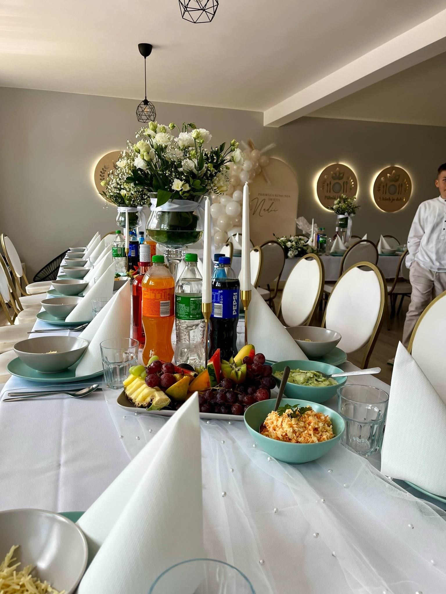 Uroczysty stół komunijny z białym obrusem zdobionym perłami, kwiatami, napojami i sałatkami. W tle dekoracje z balonów i podświetlane napisy.