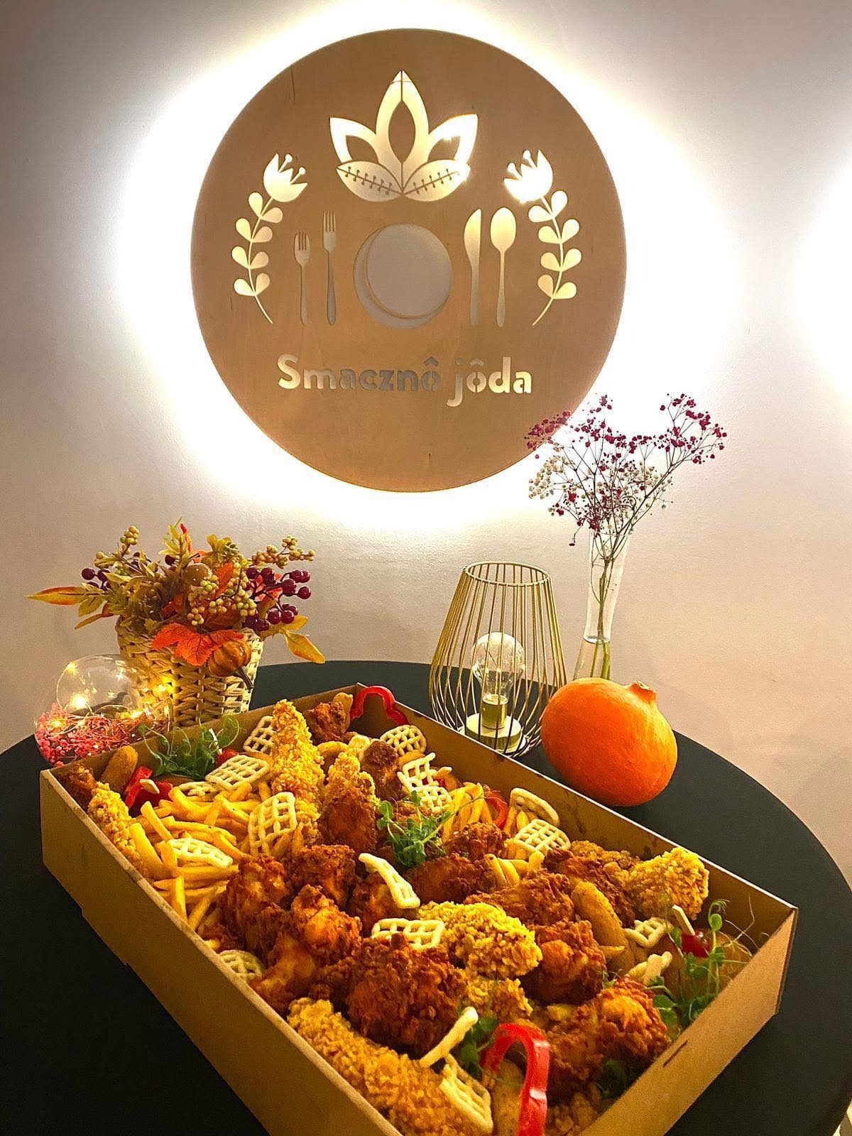 Dekoracyjna skrzynia z frytkami, kurczakiem i goframi, podświetlona logo 'Smaczna Jôda', obok dynia i kwiaty. Elegancka prezentacja dań.