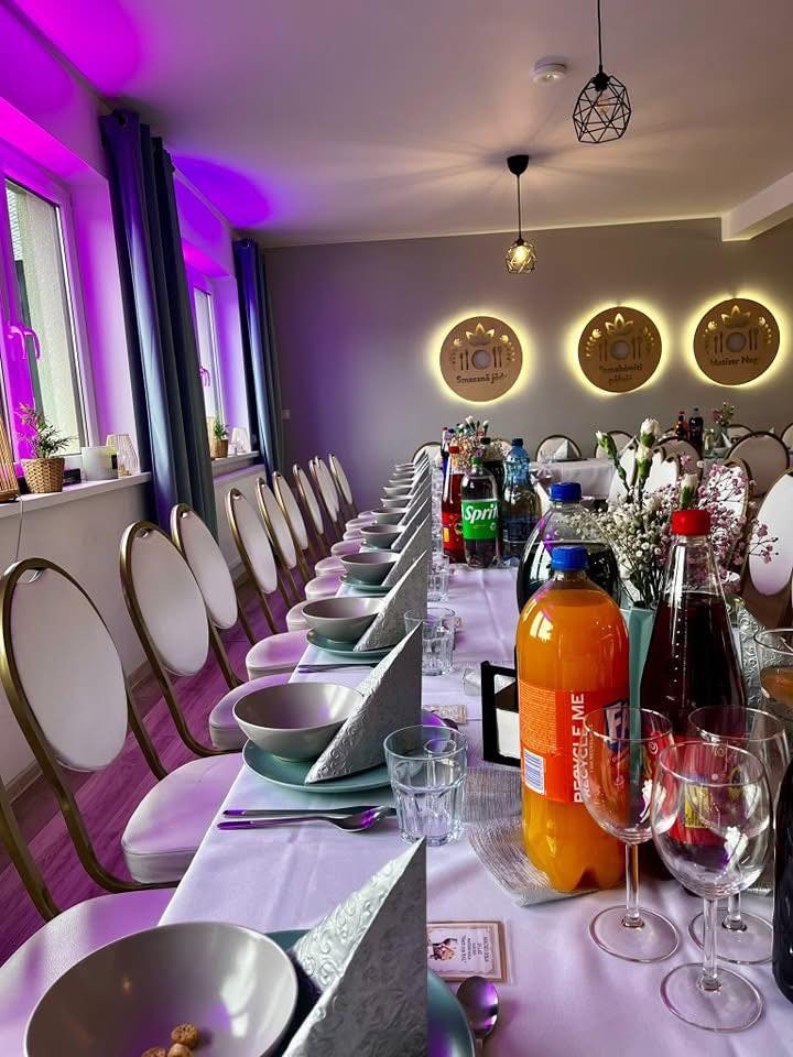 Uroczysty stół z zastawą, napojami i dekoracjami kwiatowymi, przygotowany na przyjęcie w jasnym wnętrzu z oświetleniem punktowym i fioletowymi akcentami.