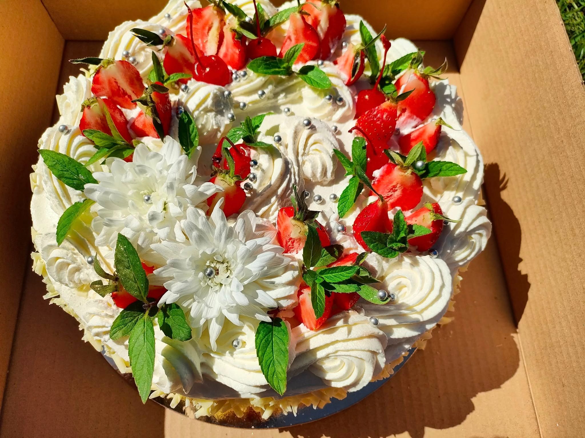 Tort śmietanowy z truskawkami, wiśniami, miętą i białymi kwiatami w kartonowym pudełku, widok z góry, oświetlenie naturalne.