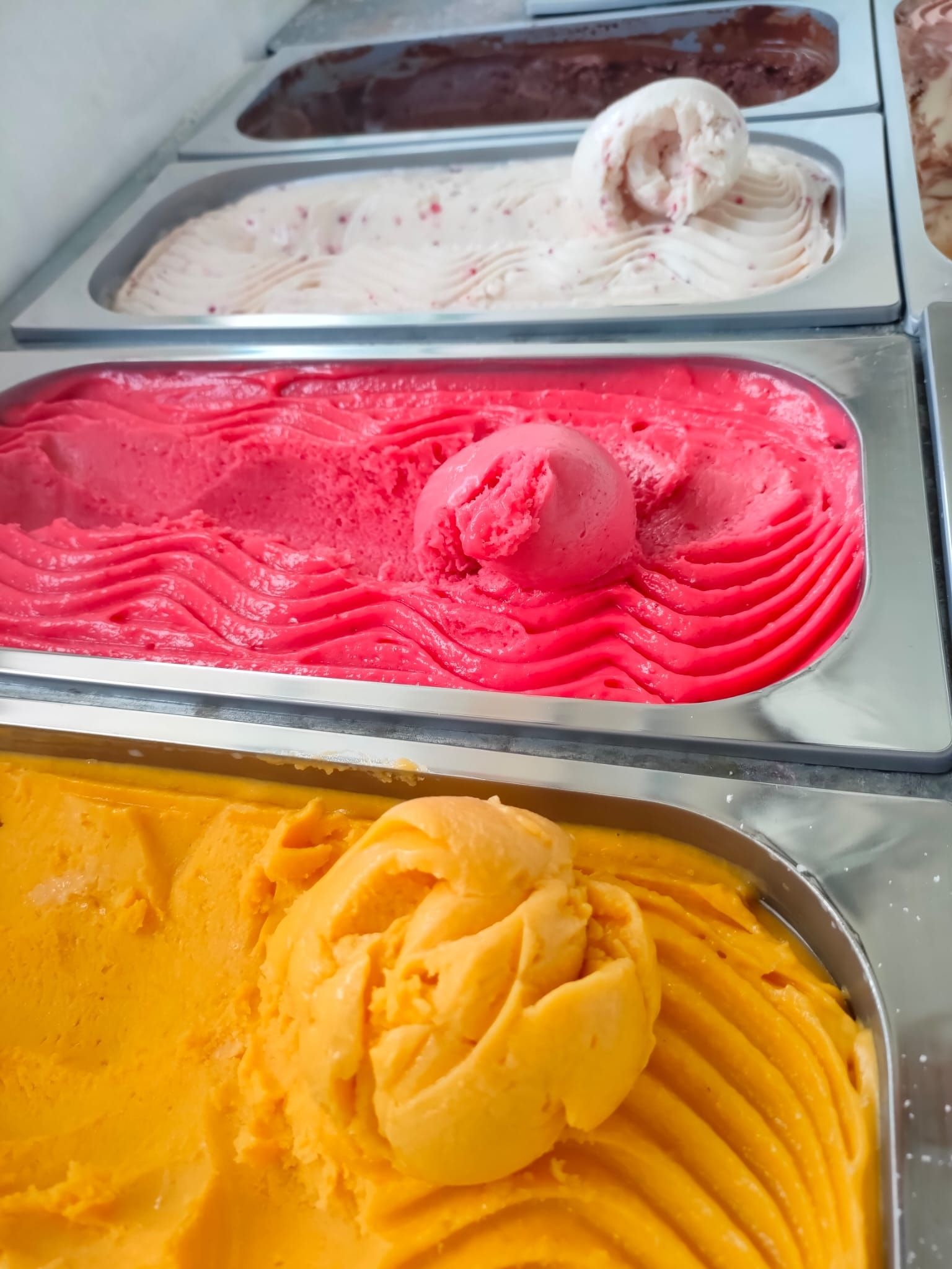 Lody w kuwetach: czekoladowe, truskawkowe, malinowe i mango. Gałki lodów ułożone w faliste wzory. Intensywne kolory lodów.