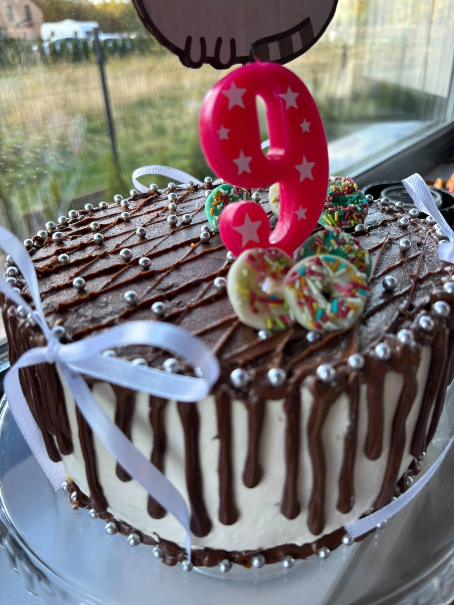 Udekorowany tort urodzinowy z polewą czekoladową, posypką z kulek i świeczką w kształcie cyfry 9, ozdobiony wstążką i mini-pączkami.