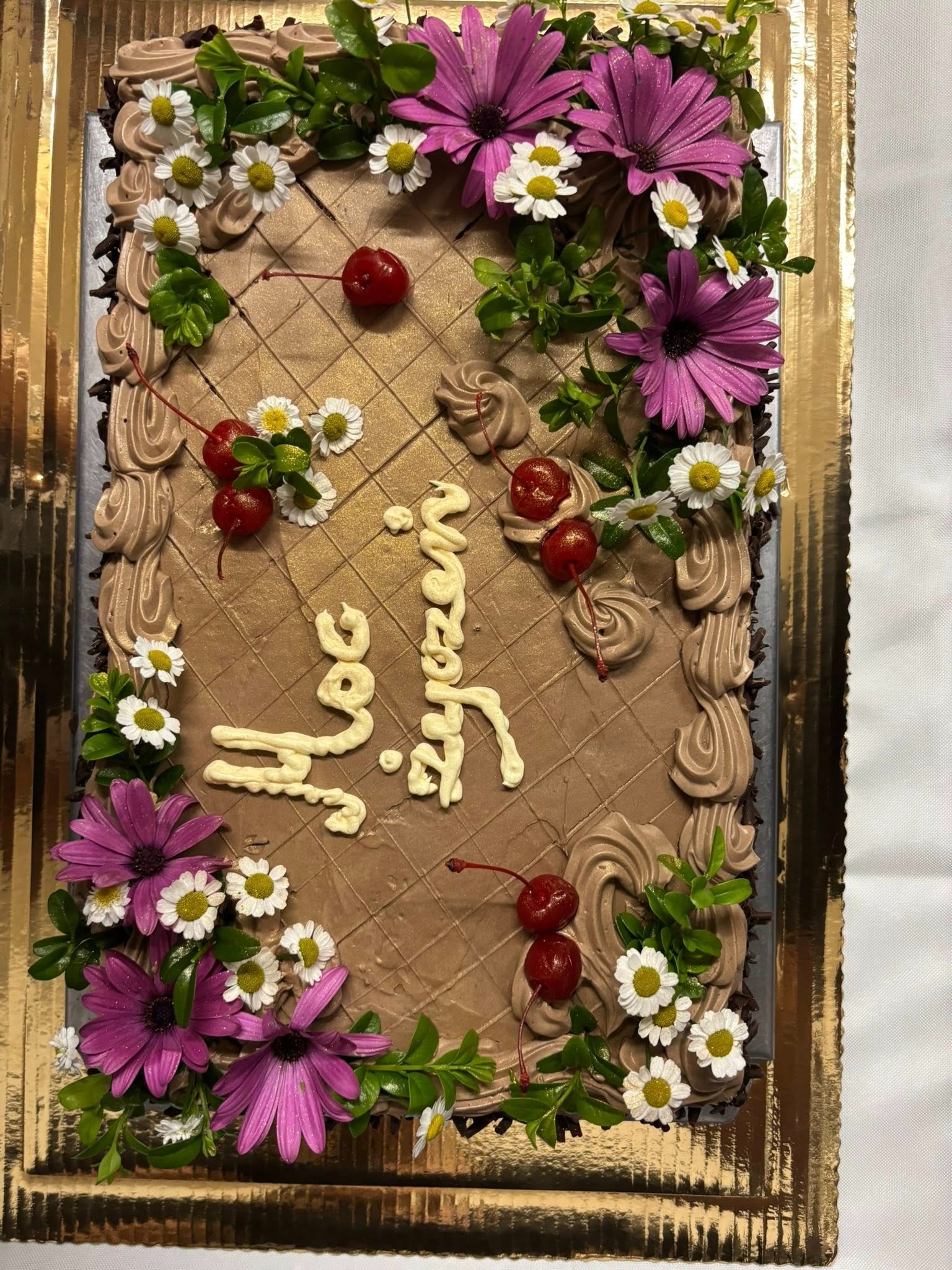 Prostokątny tort czekoladowy z kremem, wiśniami i kwiatami, na złotej tacy. Napis z kremu 'Sto lat' na wierzchu. Dekoracja z kremowych rozetek na obrzeżach.