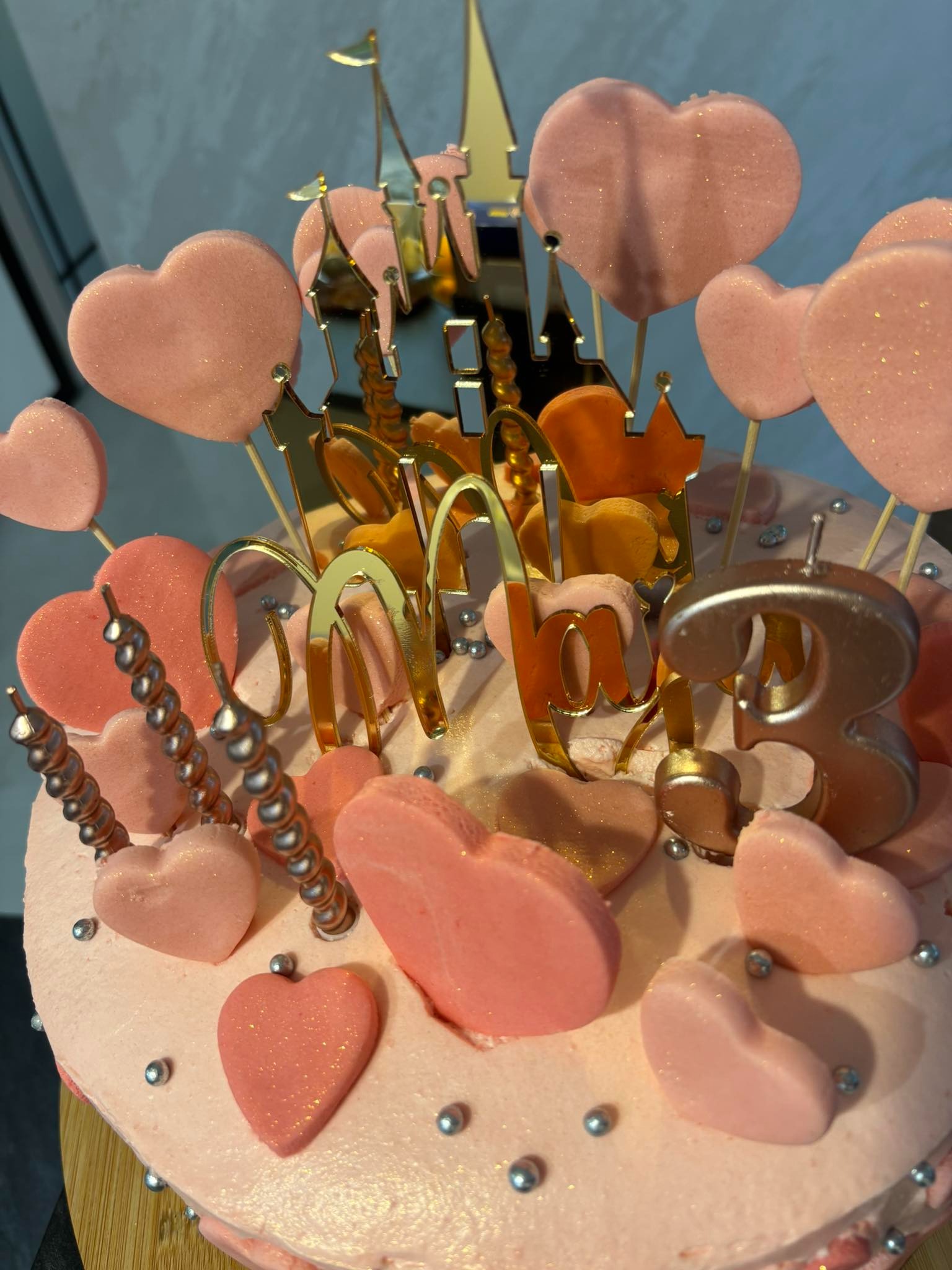 Różowy tort urodzinowy z dekoracjami w kształcie serc, zamku i cyfry 3, ozdobiony srebrnymi kuleczkami. Złote napisy i świeczki dodają blasku.