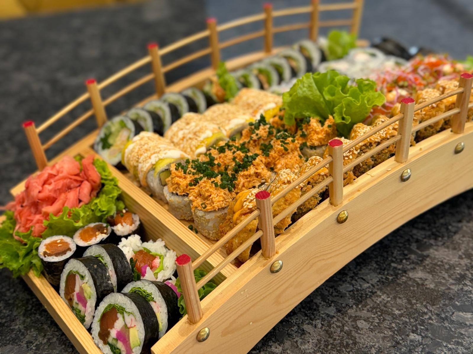 Zestaw sushi, różnorodne rolki ułożone na drewnianej tacy w kształcie mostu, z dekoracjami z imbiru i sałaty, prezentacja dań kuchni japońskiej.