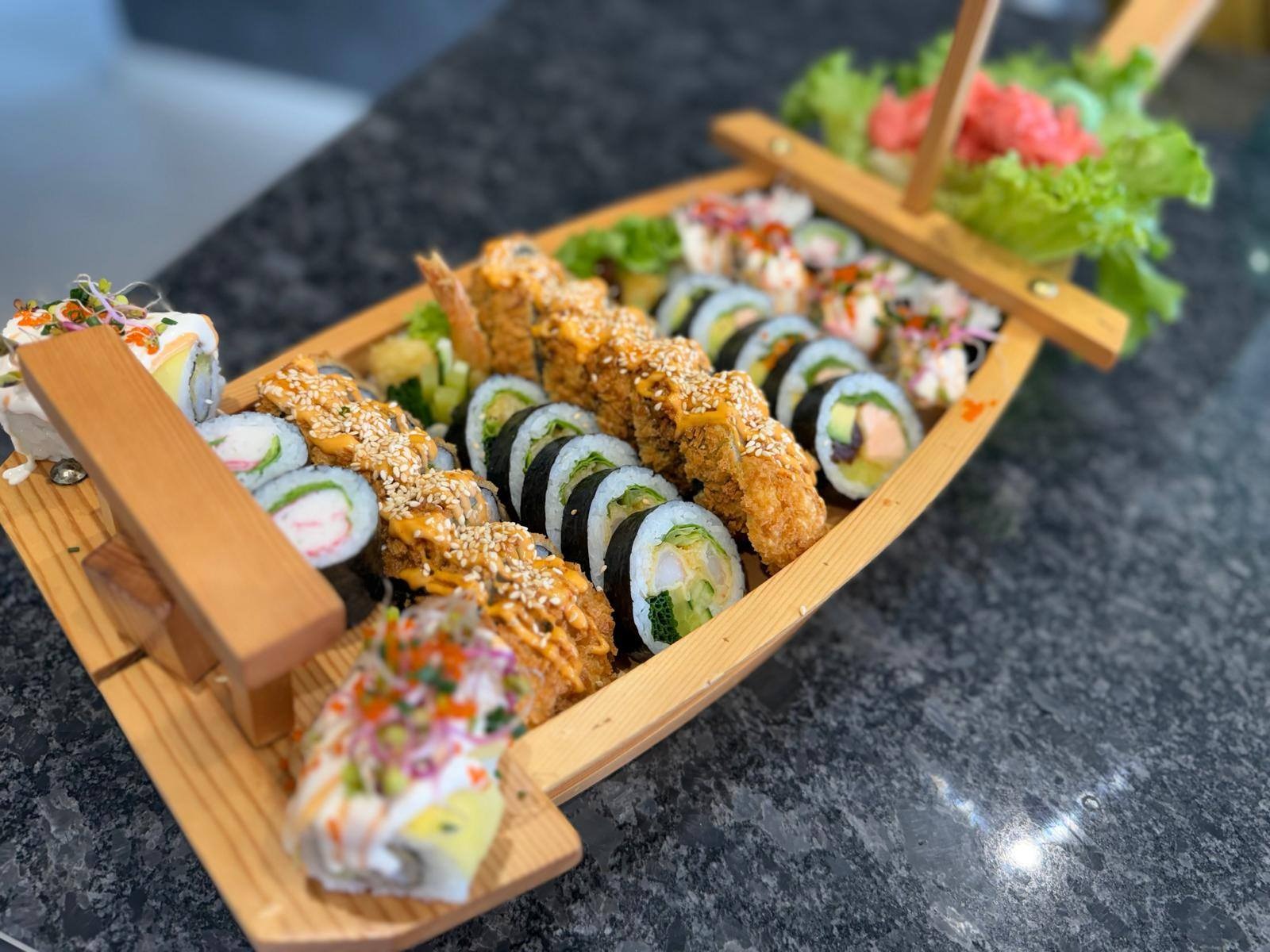 Zestaw sushi w drewnianej łódce: różnorodne rolki z krewetkami w tempurze, łososiem i warzywami, udekorowane sosem i sezamem, podane na ciemnym blacie.