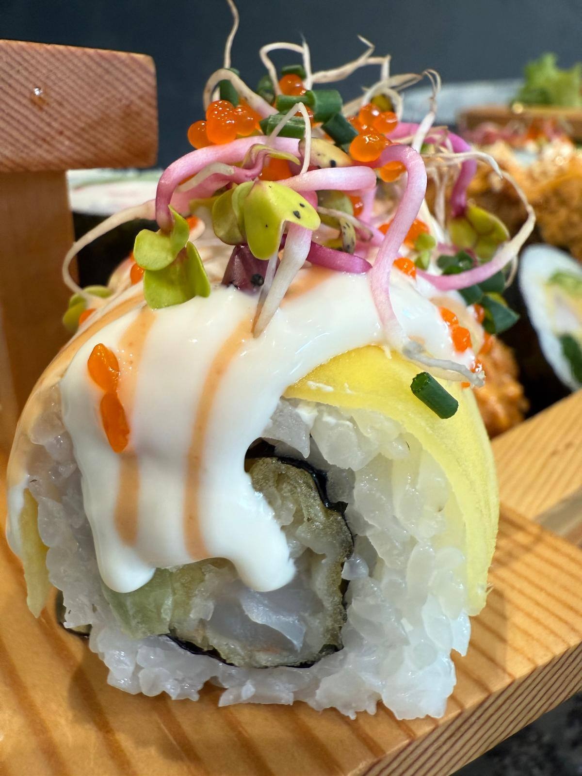 Sushi rolka z omletem, kremowym sosem i kiełkami, podana na drewnianej desce. Widoczne inne rolki w tle. Ujęcie z bliska, podkreślające detale i świeżość składników.