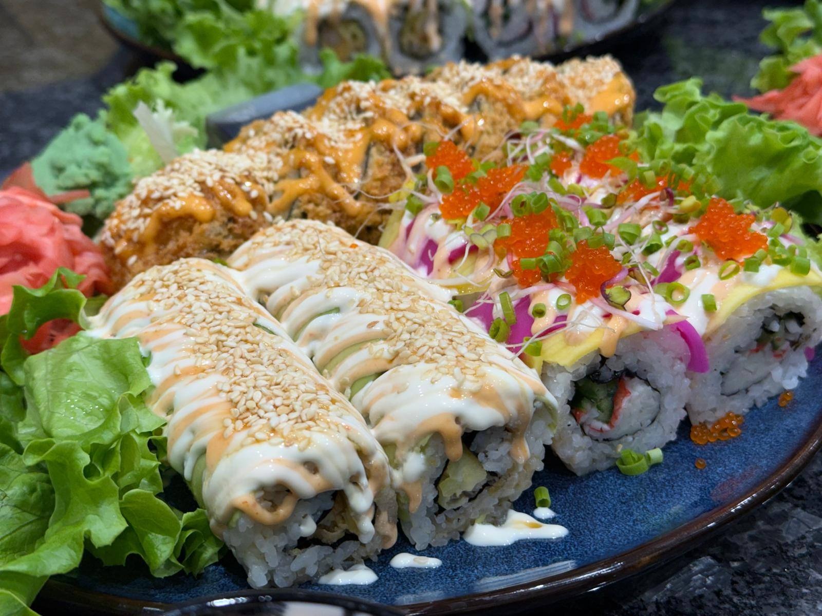 Dekoracyjne sushi z różnymi dodatkami: sezamem, sosem, kawiorem i warzywami, ułożone na niebieskim talerzu z sałatą i imbirem.