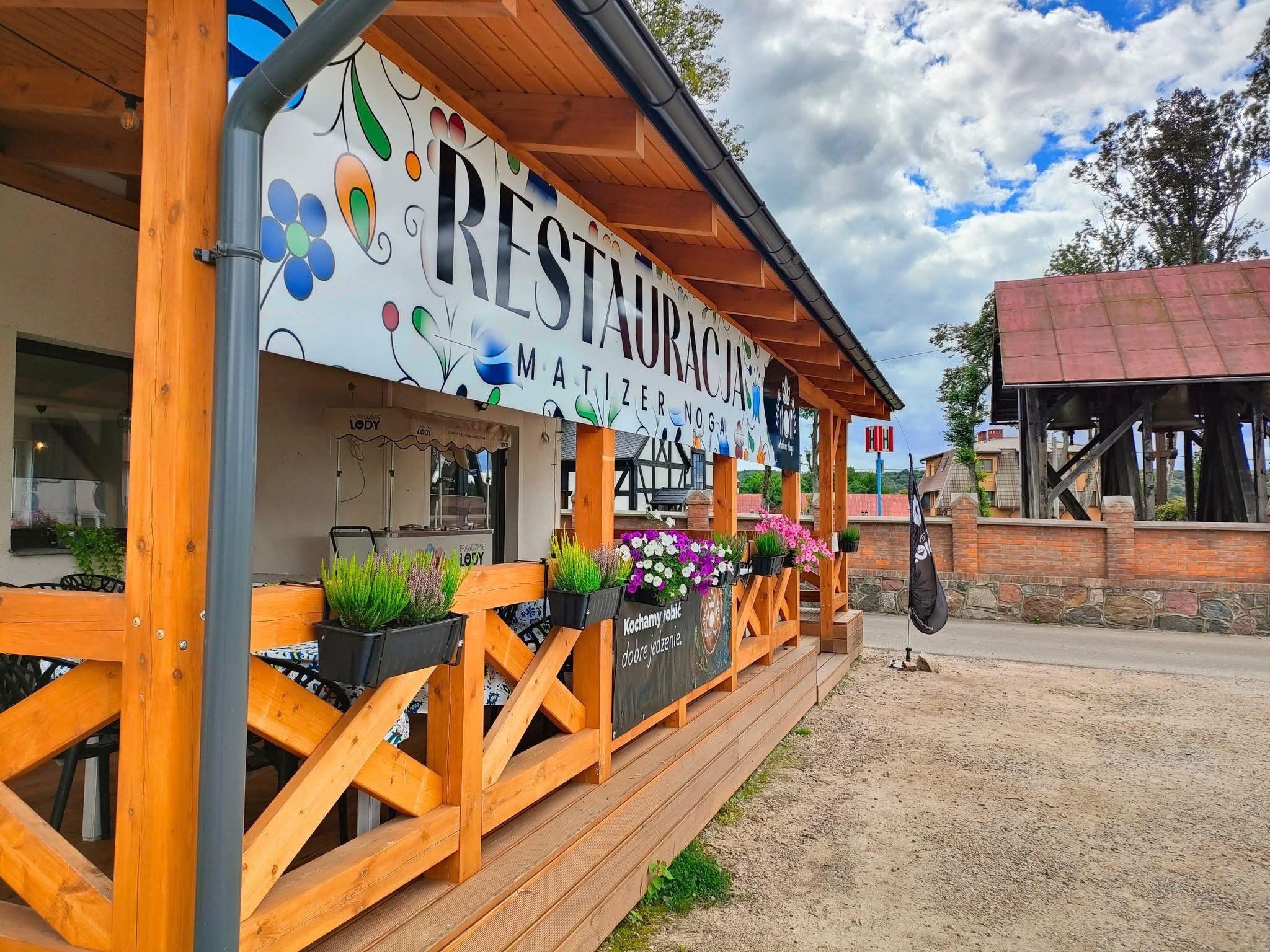 Zewnętrzny widok restauracji z drewnianym tarasem, ozdobionym kwiatami w donicach i szyldem z nazwą. W tle widoczny budynek z czerwoną dachówką i pochmurne niebo.