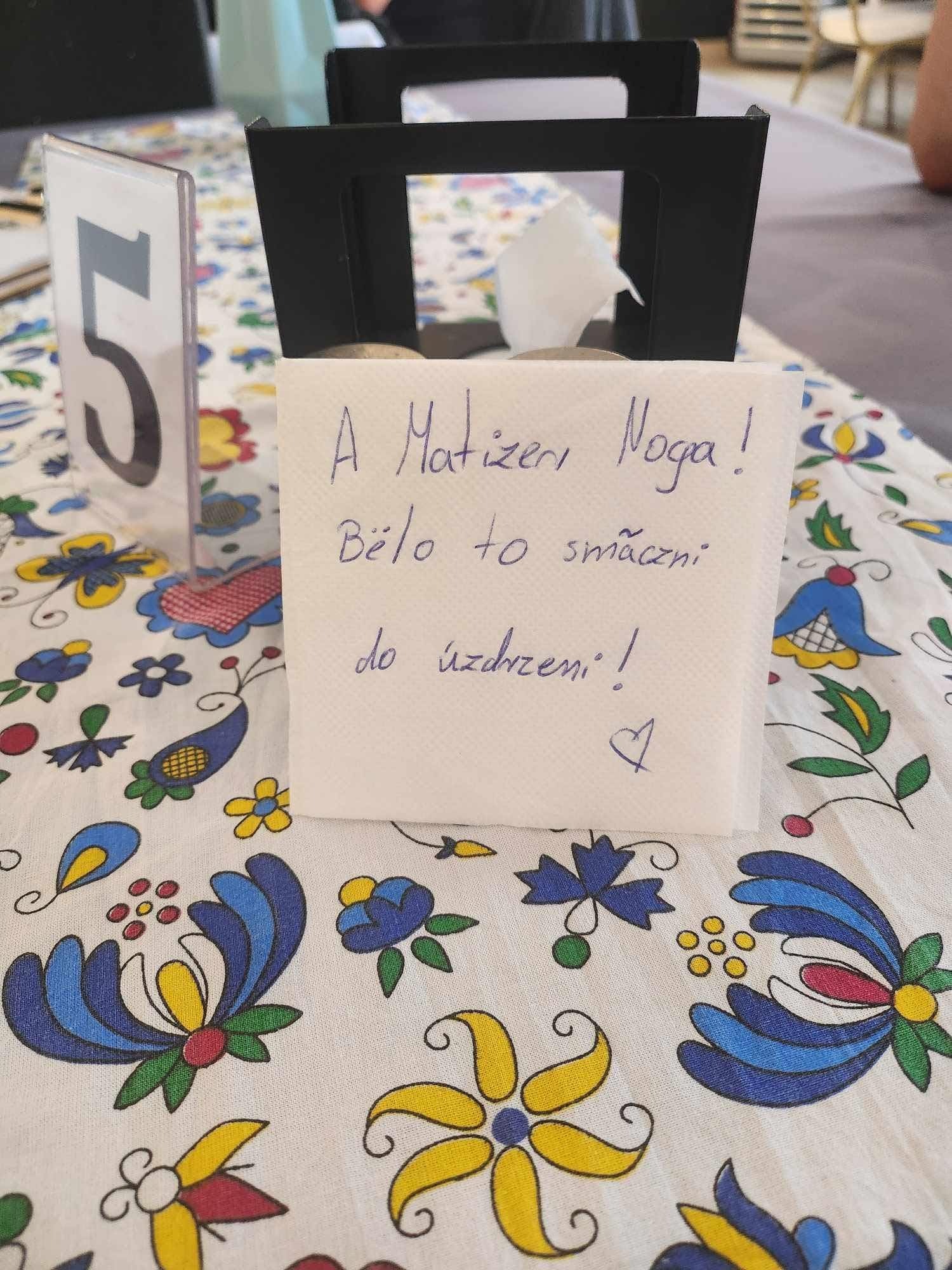 Stolik z numerem '5' i serwetką z odręcznym podziękowaniem na kwiecistym obrusie. Ujęcie z bliska, podkreślające osobisty charakter podziękowania.