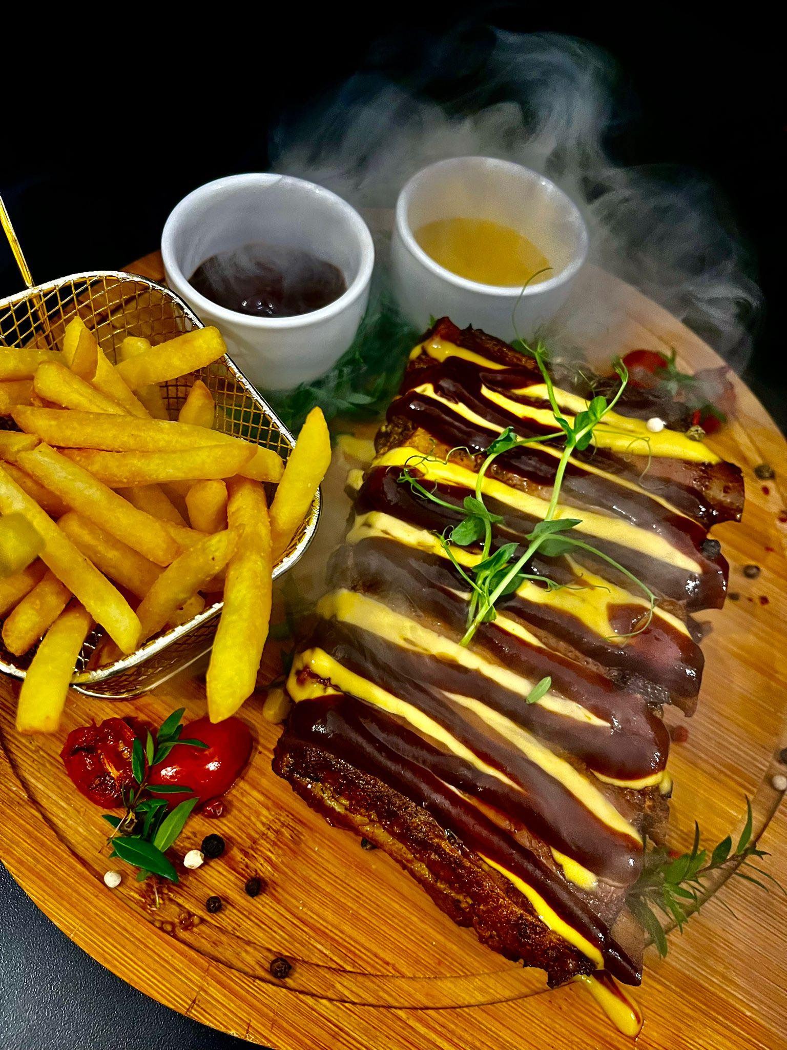Żeberka BBQ z frytkami w koszyczku, podane na drewnianej desce z sosem i dekoracją z ziół, otoczone dymem. Kulinarna kompozycja w ciemnej tonacji.