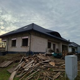 Firma Budowlana Rafał Bednarz - Niedokończony dom z ciemnym dachem i oknami bez szyb, otoczony stertą drewna i ziemią. Widoczne elementy izolacji. Pochmurne niebo w tle.