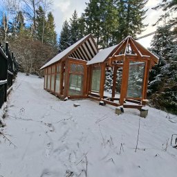 Firma Budowlana Rafał Bednarz - Drewniana konstrukcja altany/oranżerii w zimowym krajobrazie, częściowo pokryta śniegiem, z widocznym lasem w tle i fragmentem czarnego ogrodzenia po lewej stronie.