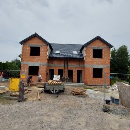 Firma Budowlana Rafał Bednarz - Wznoszony dom dwulokalowy z czerwonej cegły, z czarnym dachem i oknami bez szyb. Na pierwszym planie przyczepka z materiałami budowlanymi i pracownicy budowlani.