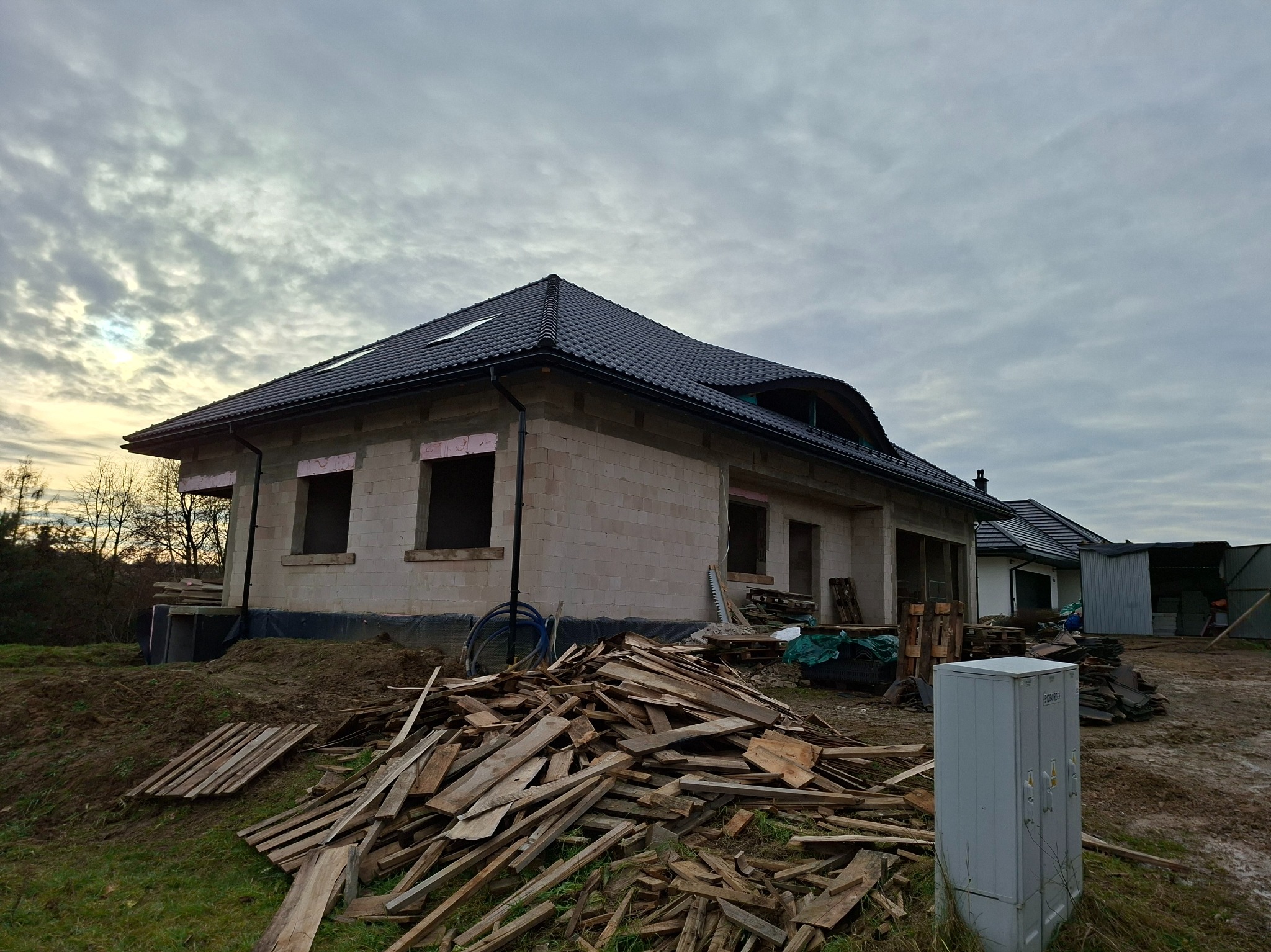 Niedokończony dom z ciemnym dachem i oknami bez szyb, otoczony stertą drewna i ziemią. Widoczne elementy izolacji. Pochmurne niebo w tle.