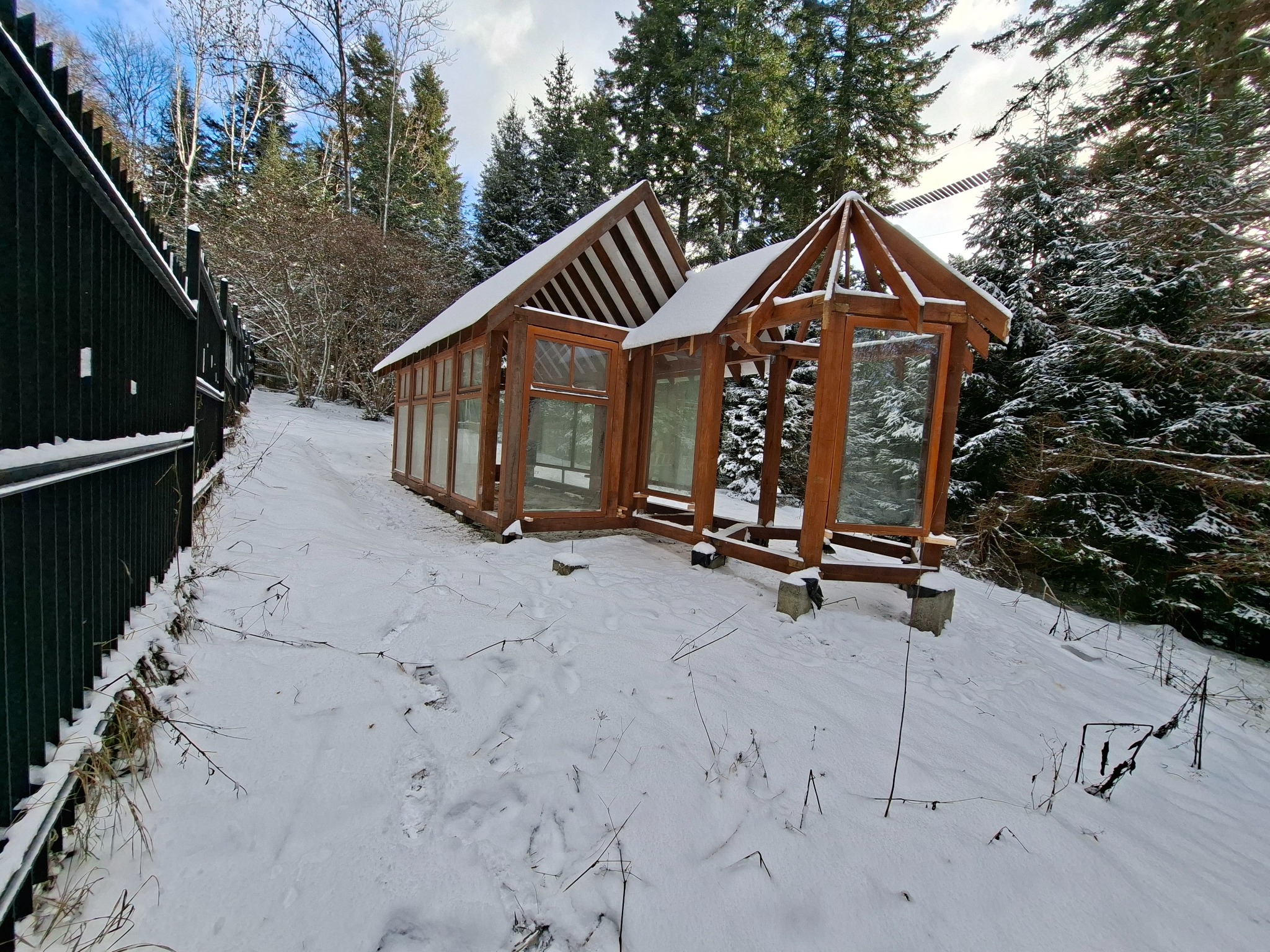 Drewniana konstrukcja altany/oranżerii w zimowym krajobrazie, częściowo pokryta śniegiem, z widocznym lasem w tle i fragmentem czarnego ogrodzenia po lewej stronie.