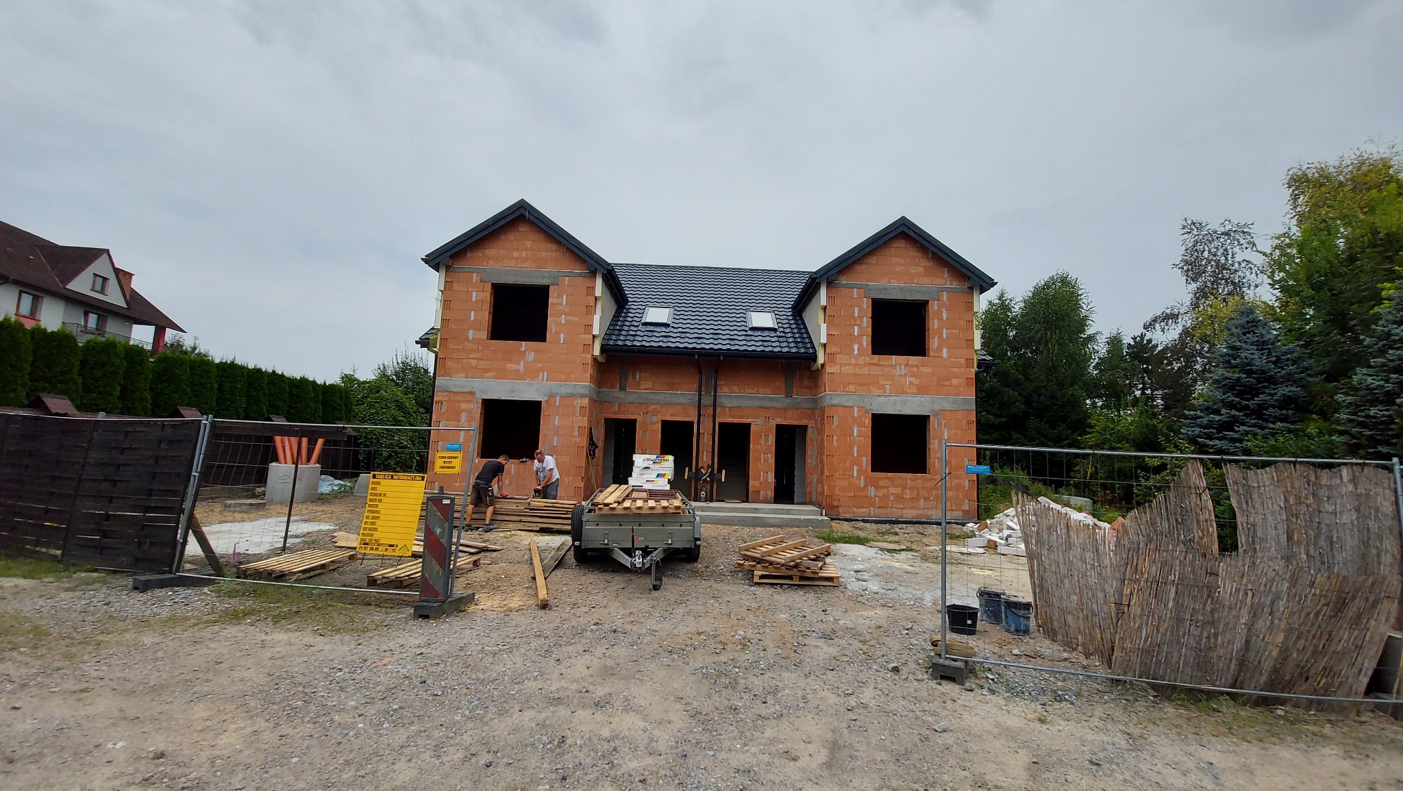 Wznoszony dom dwulokalowy z czerwonej cegły, z czarnym dachem i oknami bez szyb. Na pierwszym planie przyczepka z materiałami budowlanymi i pracownicy budowlani.