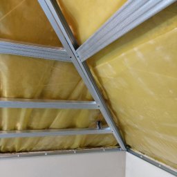 DDD FORTE Tomasz Wesołowski - Sufit podwieszany z metalową konstrukcją i izolacją z folii w kolorze żółtym, widok z dołu, kątowe ujęcie, fragment ściany w dolnej części kadru.