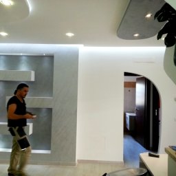 DDD FORTE Tomasz Wesołowski - Wykończone wnętrze z dekoracyjną ścianą i podwieszanym sufitem. Mężczyzna w tle, prawdopodobnie wykonawca, dodaje realizm scenie. Nowoczesny design i oświetlenie.