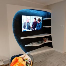 DDD FORTE Tomasz Wesołowski - Nietypowa zabudowa ściany z podświetlaną wnęką na telewizor i półkami, wykończona w kolorach bieli i czerni z efektem brokatu. Widoczne elementy wykończenia wnętrza.