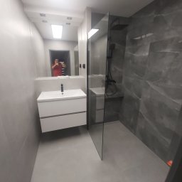 DDD FORTE Tomasz Wesołowski - Nowoczesna łazienka po remoncie: szare płytki imitujące marmur, kabina walk-in z czarną armaturą i biała szafka z umywalką. W lustrze odbija się osoba robiąca zdjęcie.