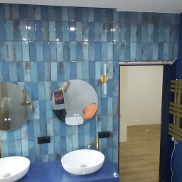 DDD FORTE Tomasz Wesołowski - Niebieska łazienka z dwoma okrągłymi lustrami, złotą armaturą i grzejnikiem. Ściany wyłożone płytkami w odcieniach błękitu i drewnianymi panelami. Nowoczesny design.