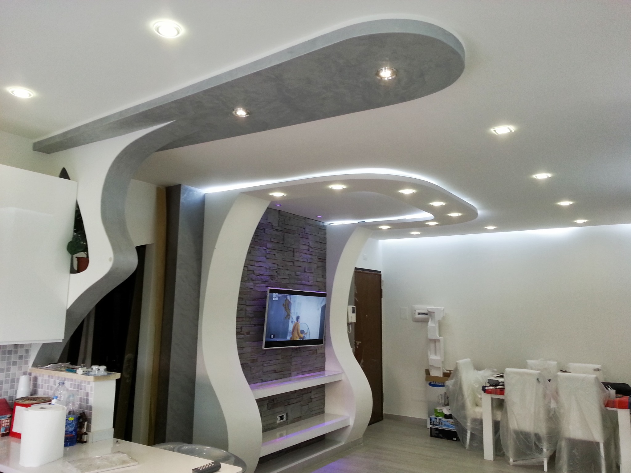 Nowoczesny salon z dekoracyjnym sufitem podwieszanym w kształcie fali, oświetleniem LED i ścianą z kamienia dekoracyjnego z telewizorem. Widoczne meble i elementy wykończenia.