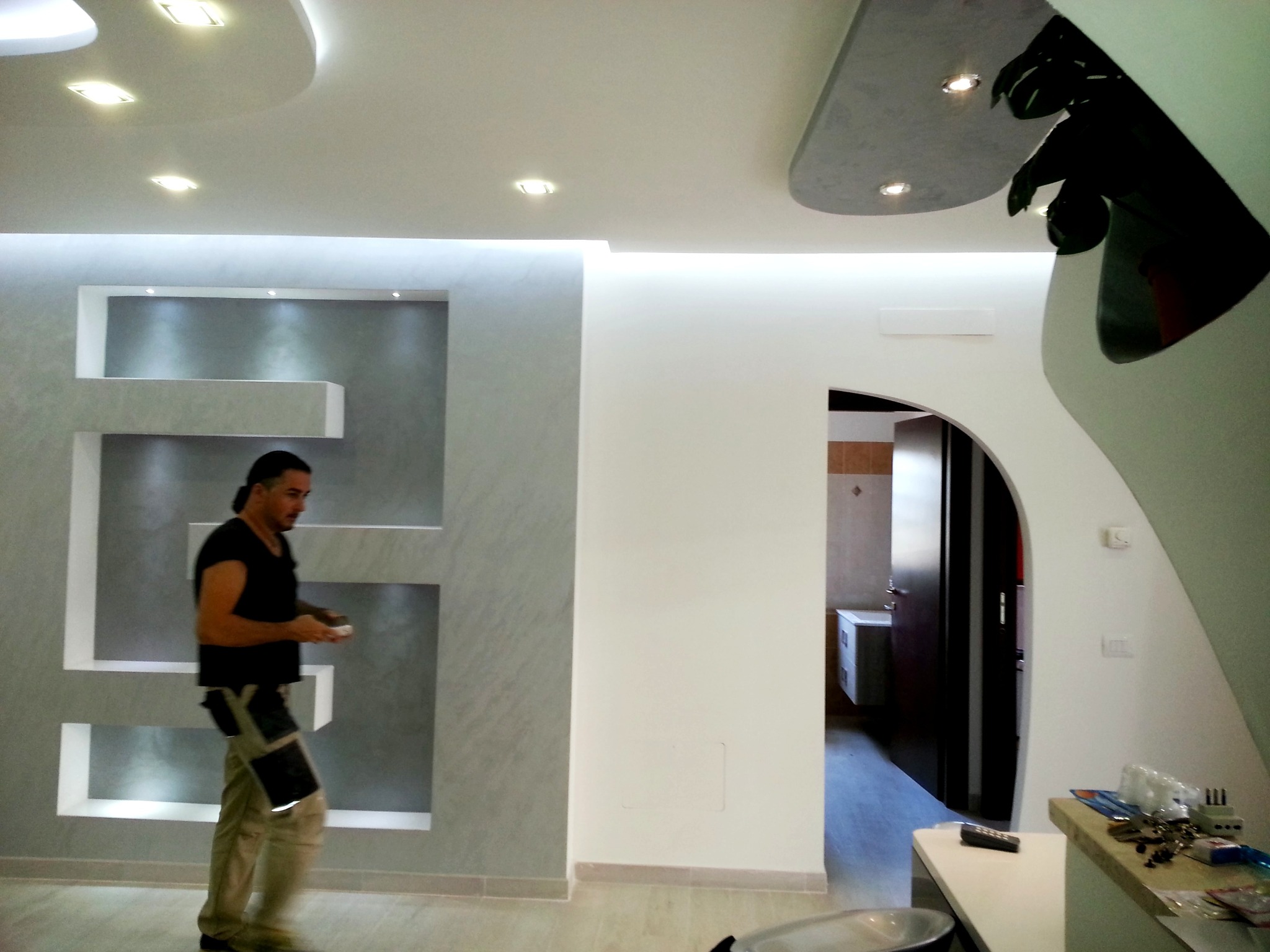 Wykończone wnętrze z dekoracyjną ścianą i podwieszanym sufitem. Mężczyzna w tle, prawdopodobnie wykonawca, dodaje realizm scenie. Nowoczesny design i oświetlenie.