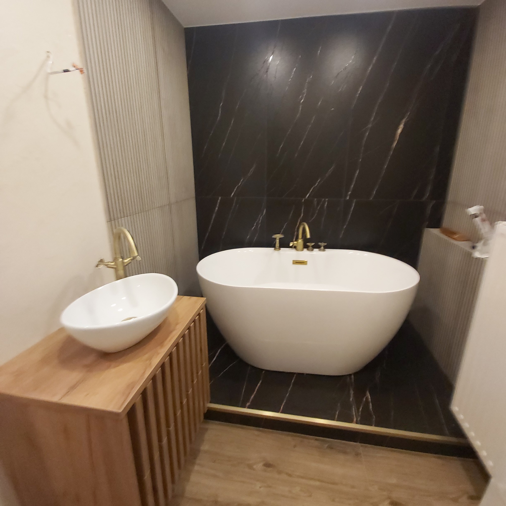 Nowoczesna łazienka z wolnostojącą wanną i umywalką nablatową na drewnianej szafce. Czarna ściana z marmurowym wzorem i złota armatura dodają elegancji.