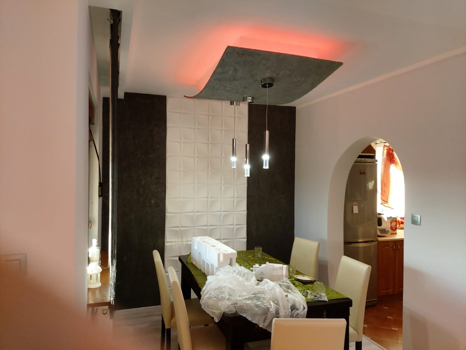 Nowoczesna jadalnia z panelami 3D na ścianie i podwieszanym sufitem z oświetleniem LED. Widoczne elementy dekoracyjne i stół z krzesłami. W tle widoczny fragment kuchni.