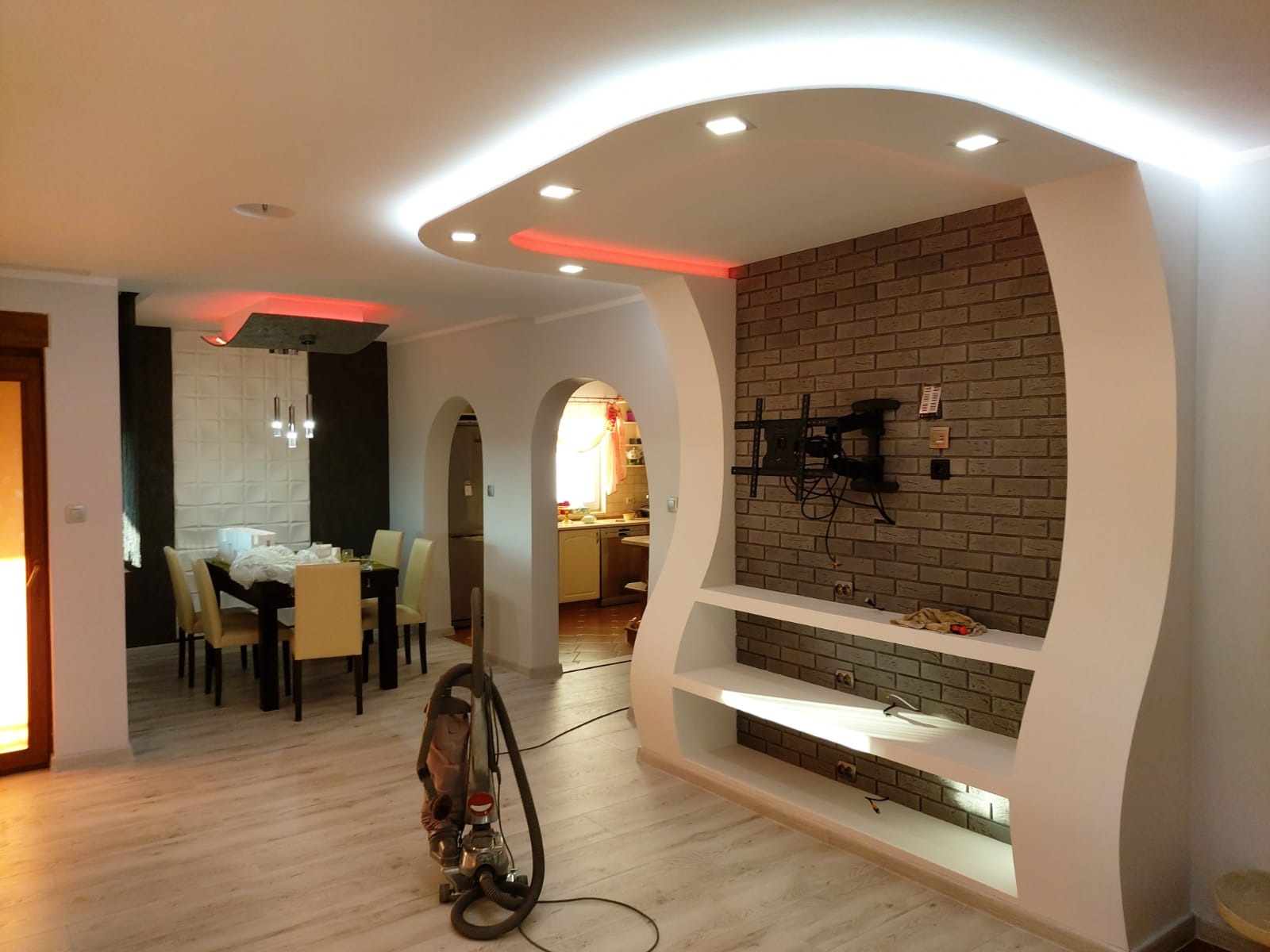 Salon po remoncie: ceglana ściana z półkami i uchwytem TV, podwieszany sufit z oświetleniem LED. Widoczny stół z krzesłami i odkurzacz. Wykończenie wnętrz.